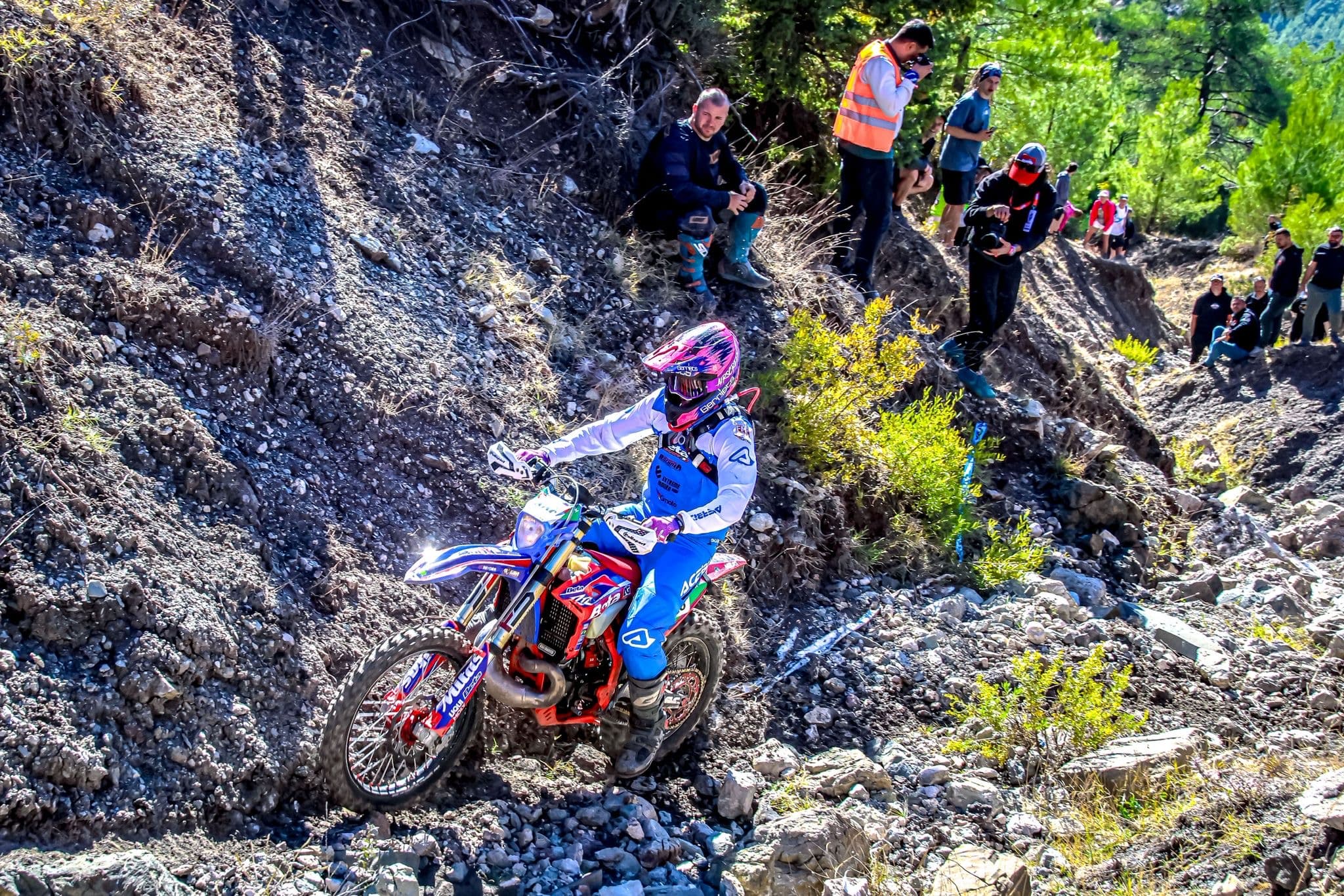 Enduro-Fahrer auf rotem Motorrad navigiert durch steiles, felsiges Gelände, beobachtet von Zuschauern am Hang.