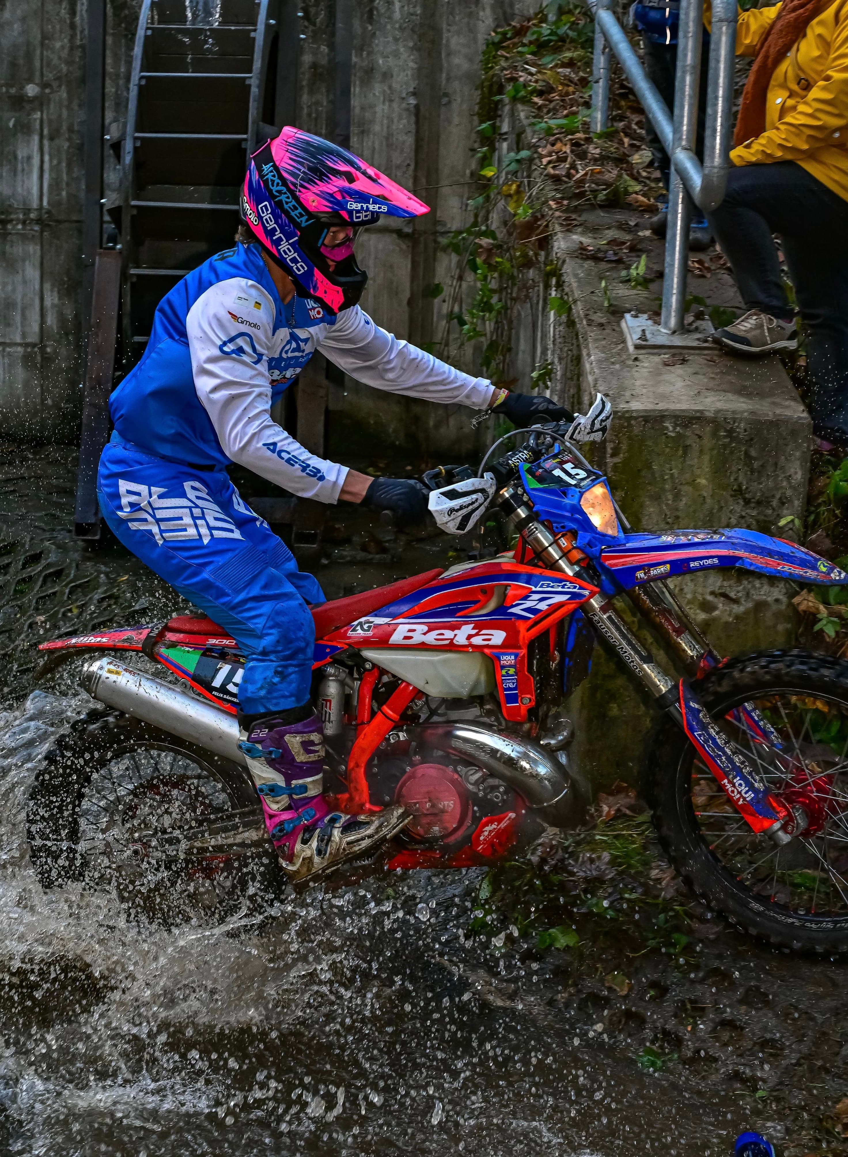 Ein Enduro-Fahrer mit pinkem Helm fährt auf einem rot-blauen Motorrad durch spritzendes Wasser.