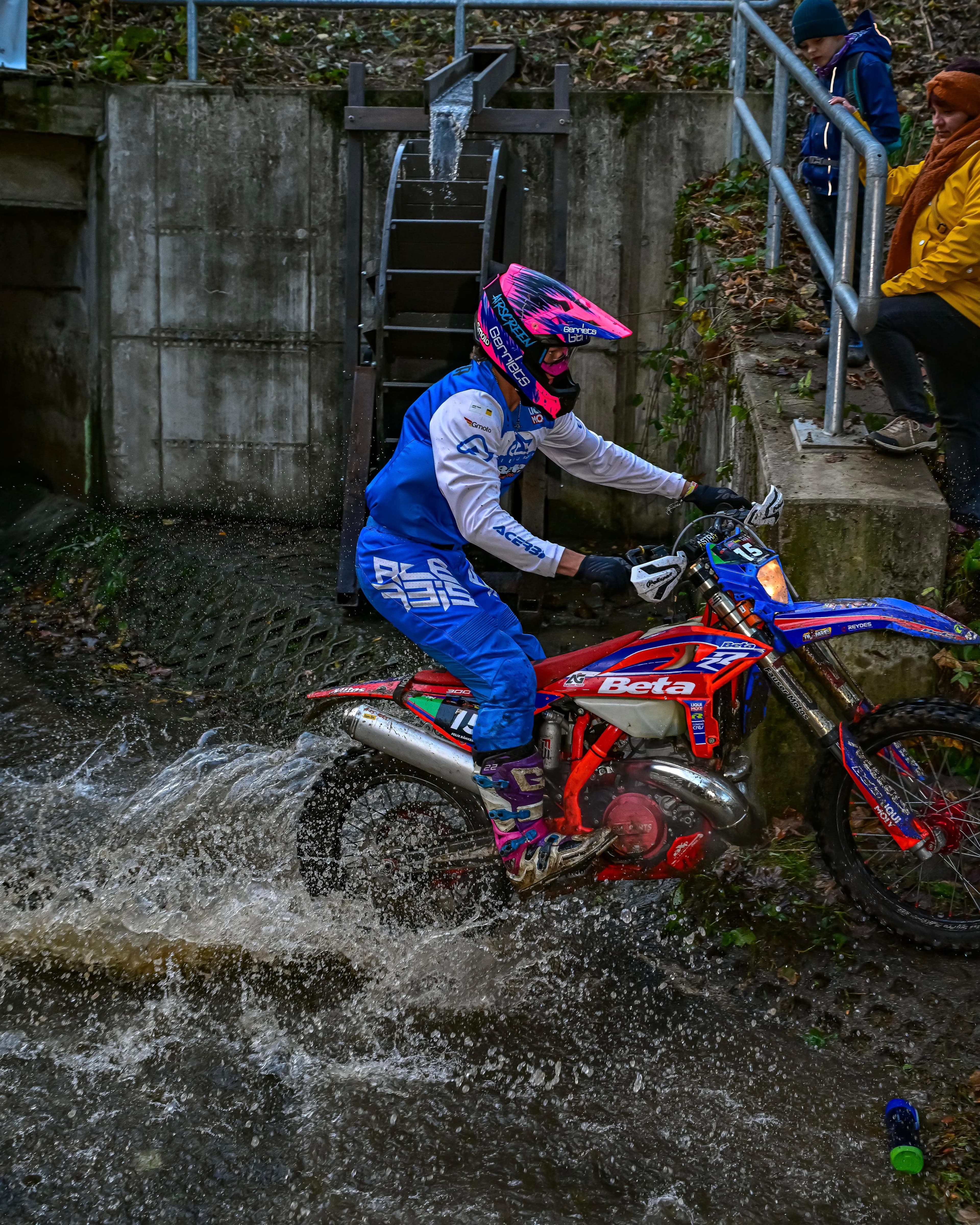 Enduro-Fahrer in blauer Kleidung fährt spritzend durch Wasser an einem Wasserrad vorbei.