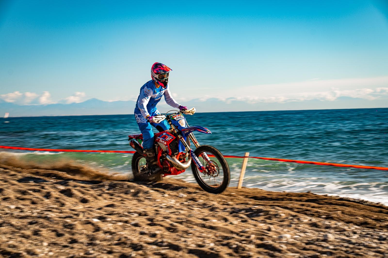 Ein Motocross-Fahrer rast auf seinem Motorrad über einen sandigen Strand am blauen Meer entlang.