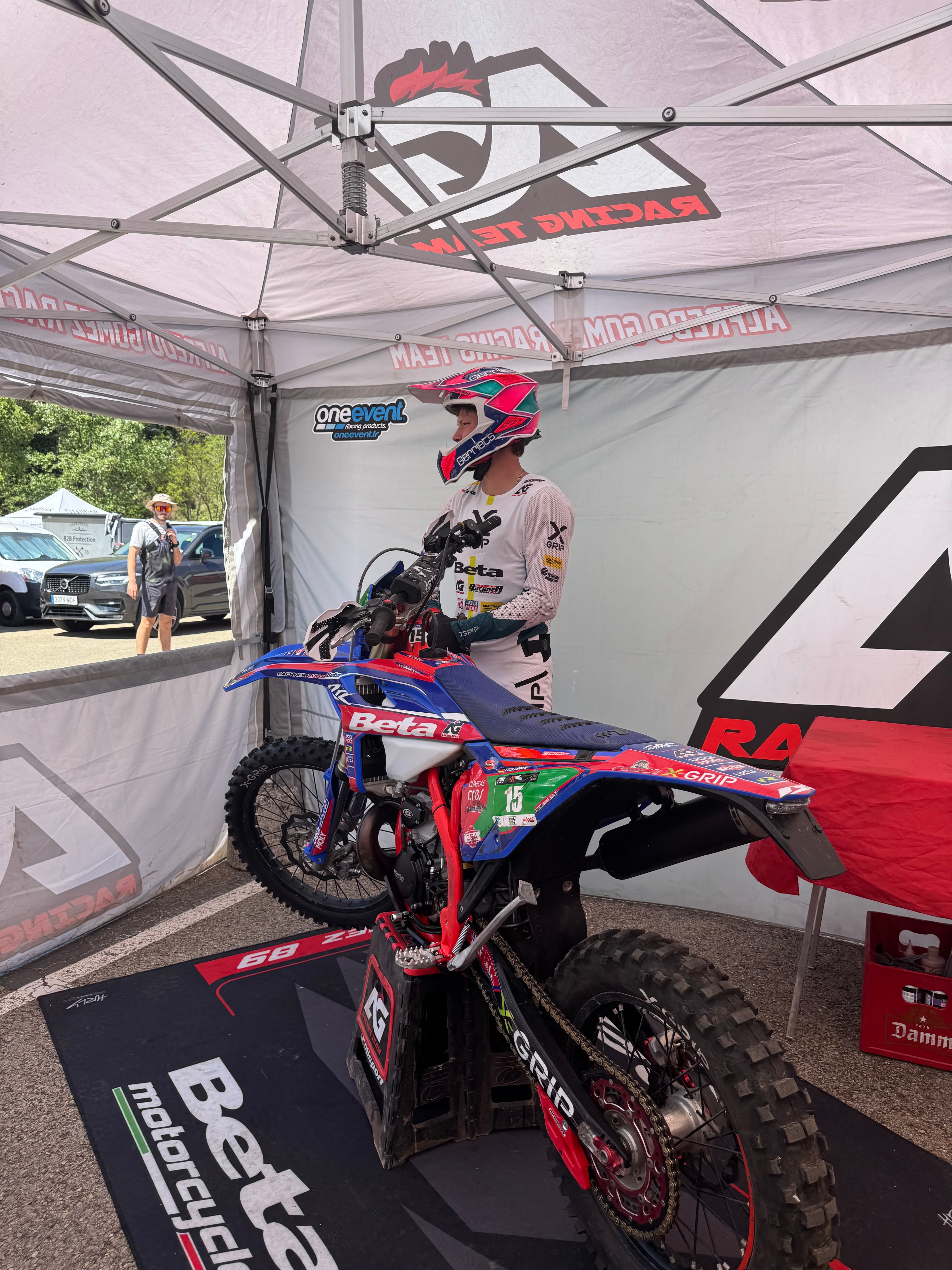 Ein Motocross-Fahrer in voller Ausrüstung steht neben seinem blau-roten Rennmotorrad unter einem weißen Zelt.