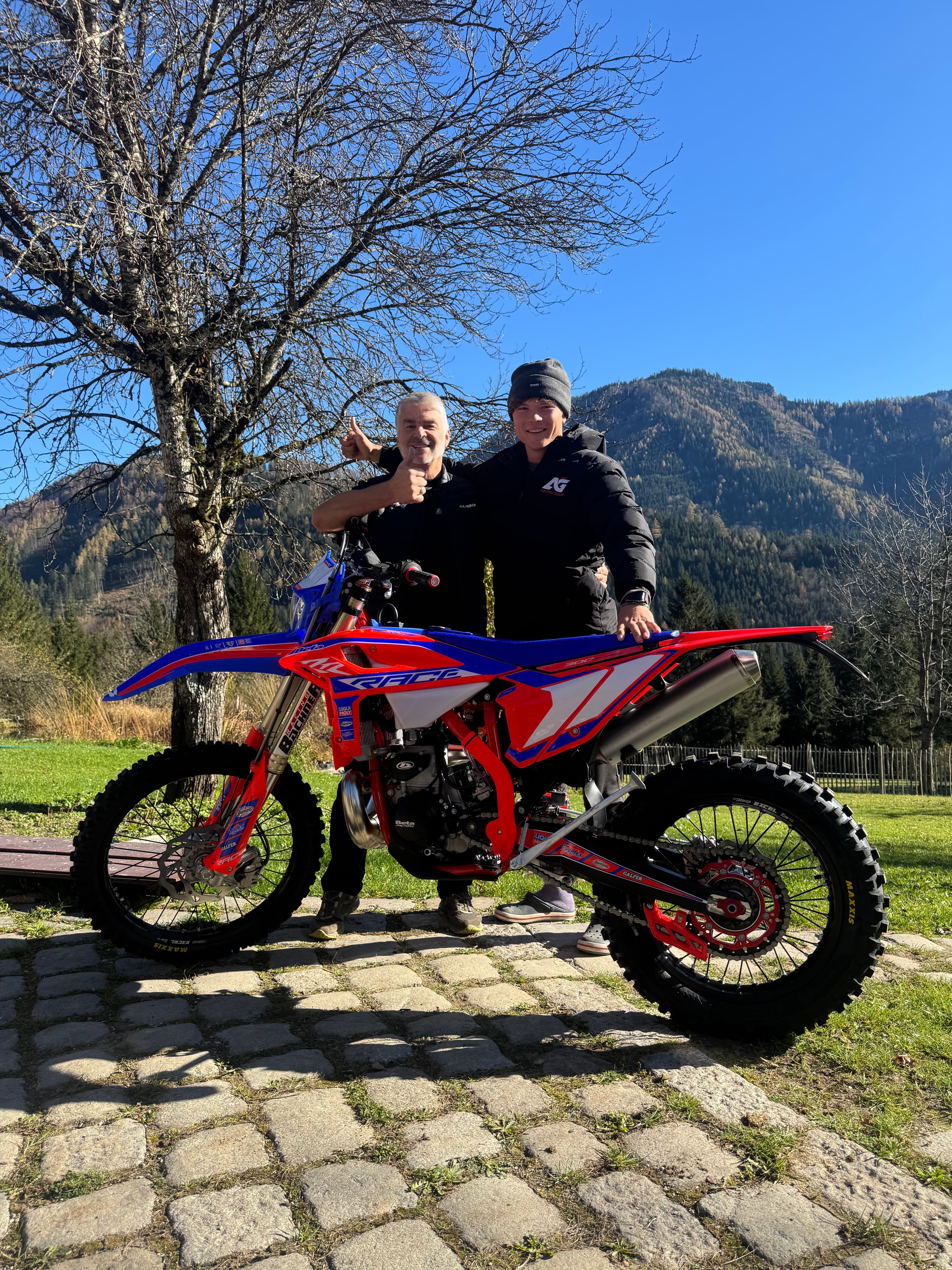 Zwei Männer posieren mit einem rot-blauen Motocross-Motorrad auf einem gepflasterten Weg vor einer Bergkulisse.