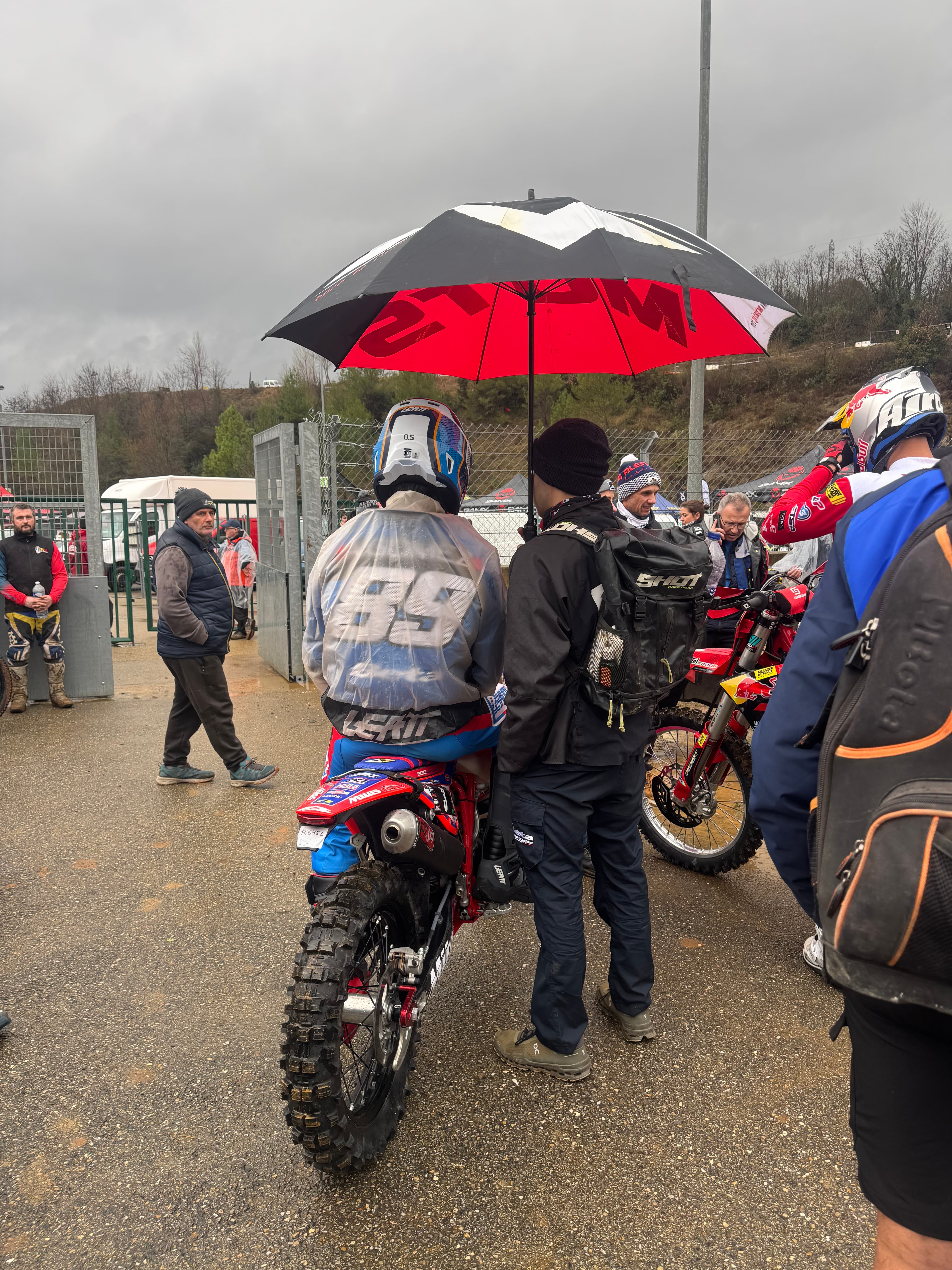 Motocross-Fahrer auf seinem Motorrad unter einem rot-schwarzen Regenschirm, umgeben von Zuschauern an der Rennstrecke.