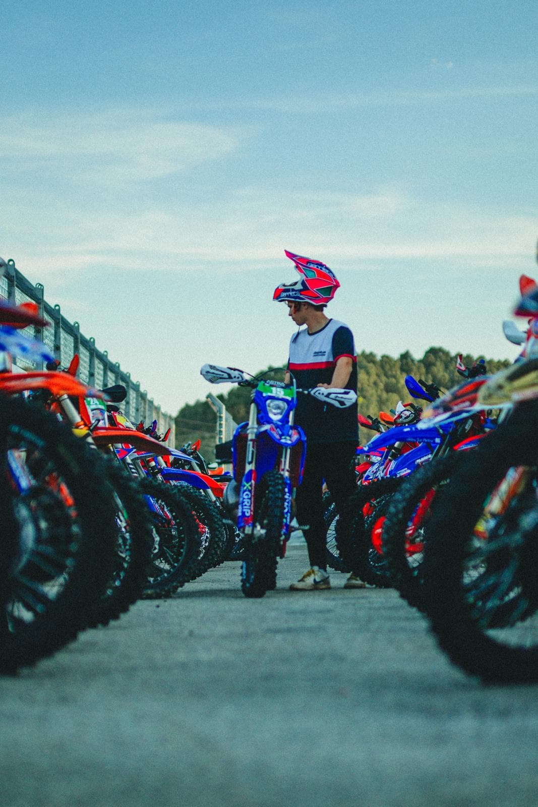 Ein Motocross-Fahrer mit pinkem Helm steht hinter einem blauen Motorrad zwischen Reihen bunter Geländemotorräder.