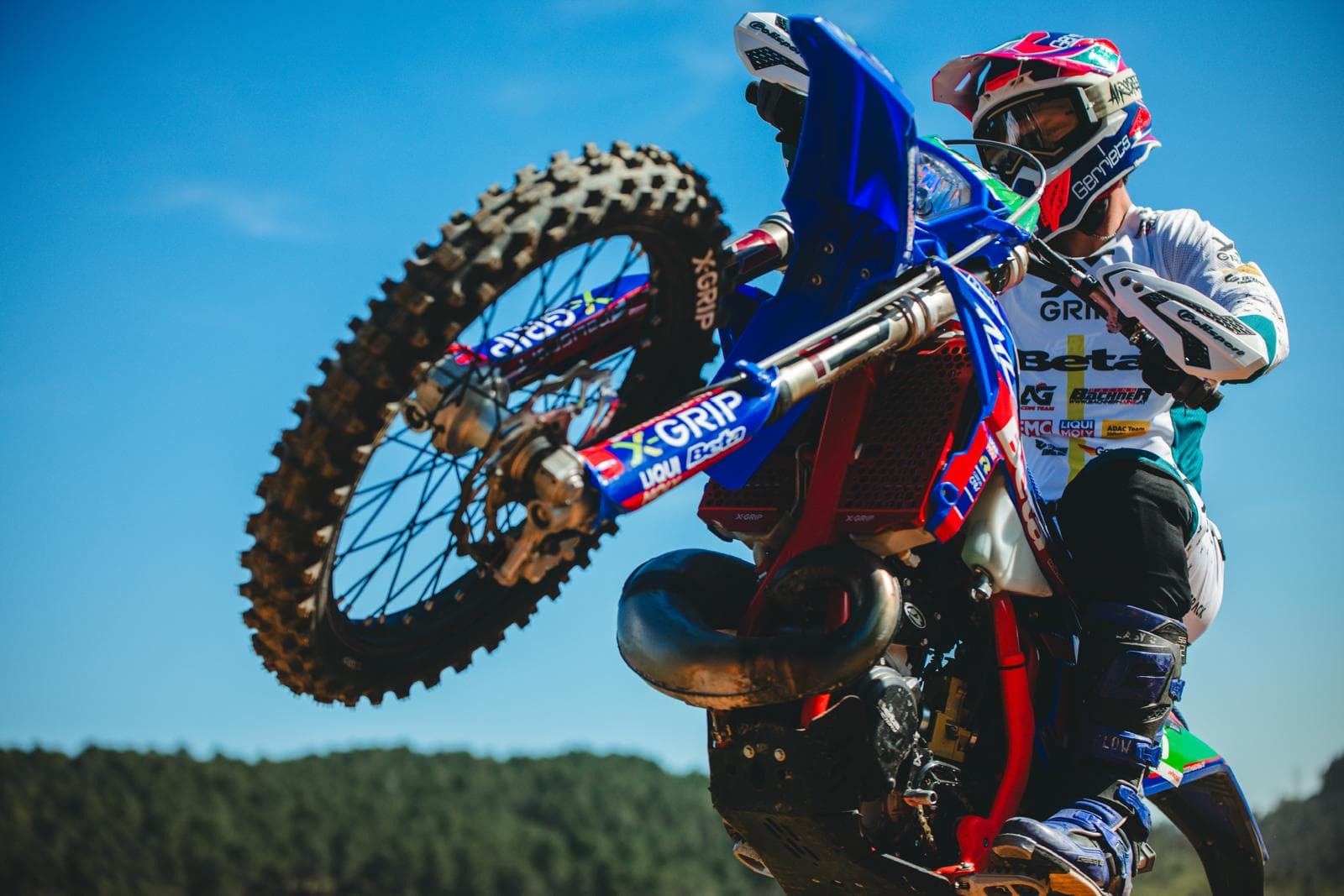 Ein Motocross-Fahrer vollführt einen Wheelie auf einer blau-roten Maschine vor strahlend blauem Himmel.
