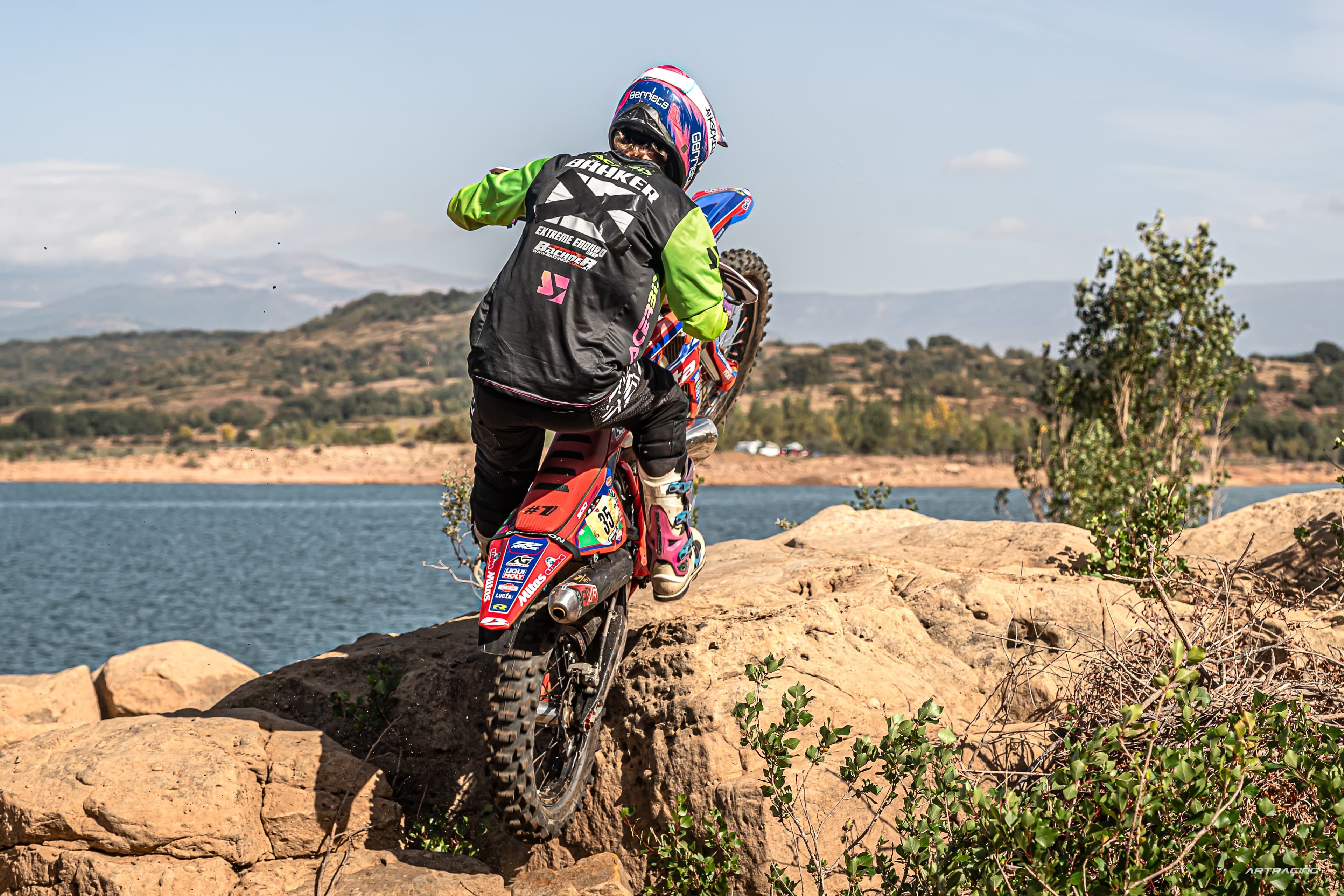 Motocross-Fahrer vollführt einen Wheelie auf felsigem Untergrund vor einem weiten See und Hügeln.