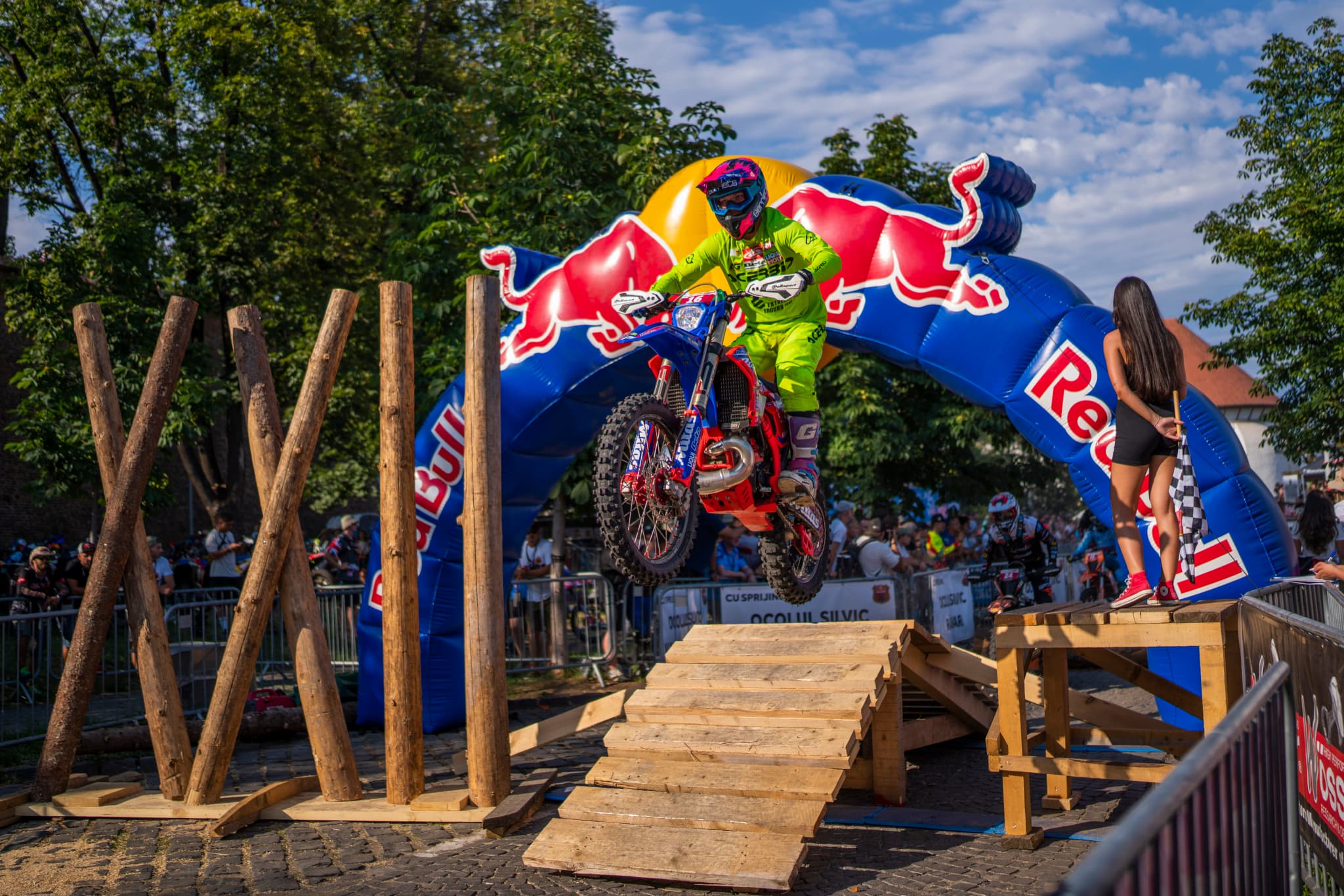 Motocross-Fahrer in neongrüner Kleidung springt über eine Holzrampe vor einem großen, aufblasbaren Red-Bull-Bogen.