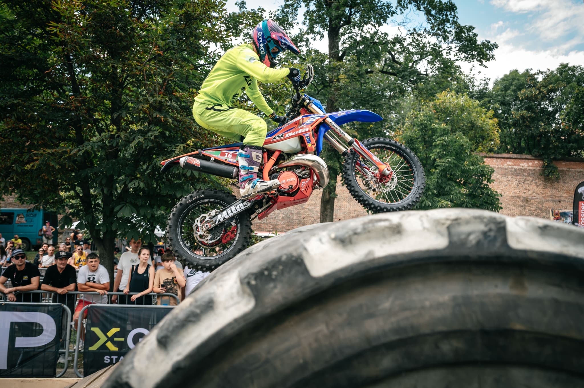 Ein Motocross-Fahrer in neongelber Kleidung überquert auf seinem Motorrad einen riesigen Traktorreifen vor Publikum.