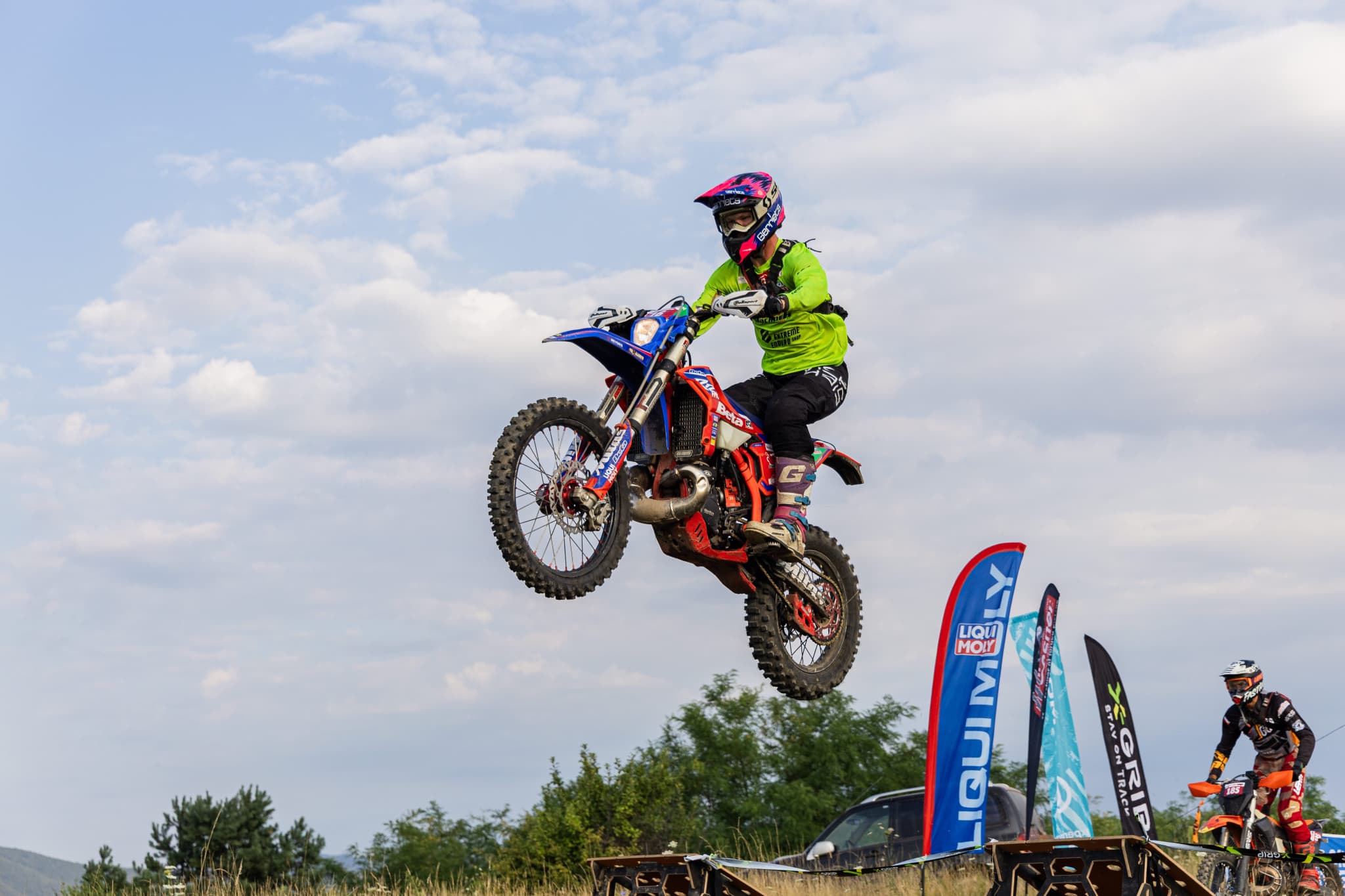 Motocross-Fahrer in neongrünem Trikot springt mit seinem blau-roten Motorrad hoch in den bewölkten Himmel.