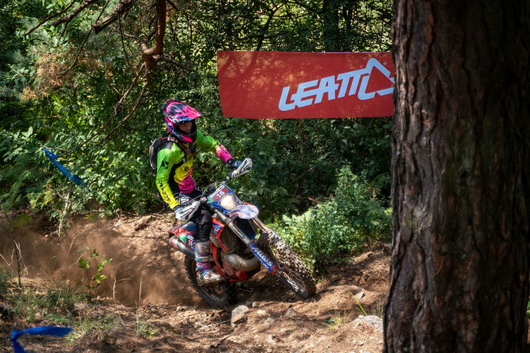 Motocross-Fahrer in bunter Ausrüstung fährt auf einem staubigen Waldpfad an einem roten Leatt-Banner vorbei.