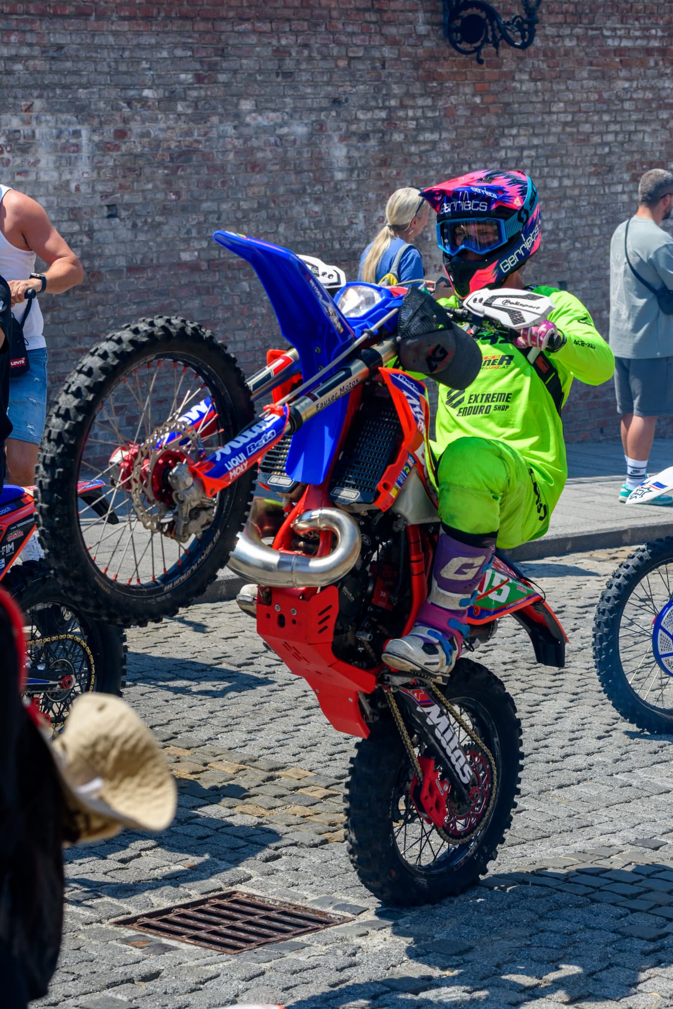 Motocross-Fahrer in neongrüner Kleidung macht einen Wheelie auf einem rot-blauen Motorrad auf Kopfsteinpflaster.